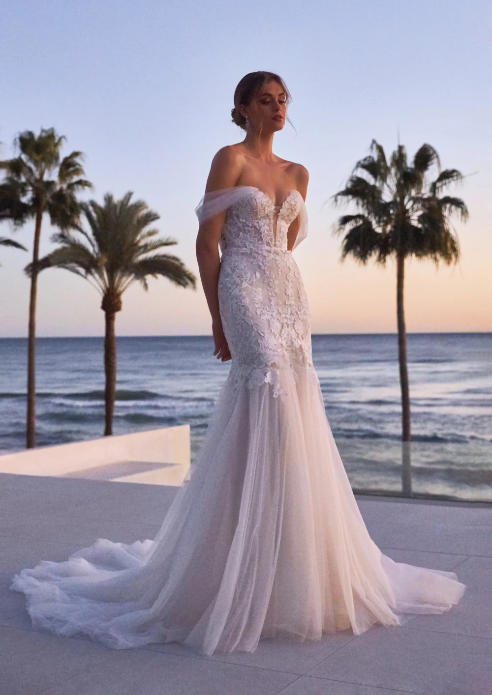 Model posing next to a beach wearing a strapless blush lace and tulle fishtail wedding dress.